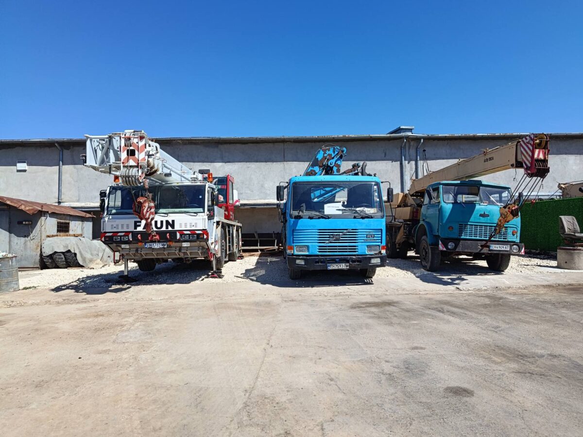 Three heavy trucks with cranes parked in a gravel lot in front of a warehouse on a sunny day.