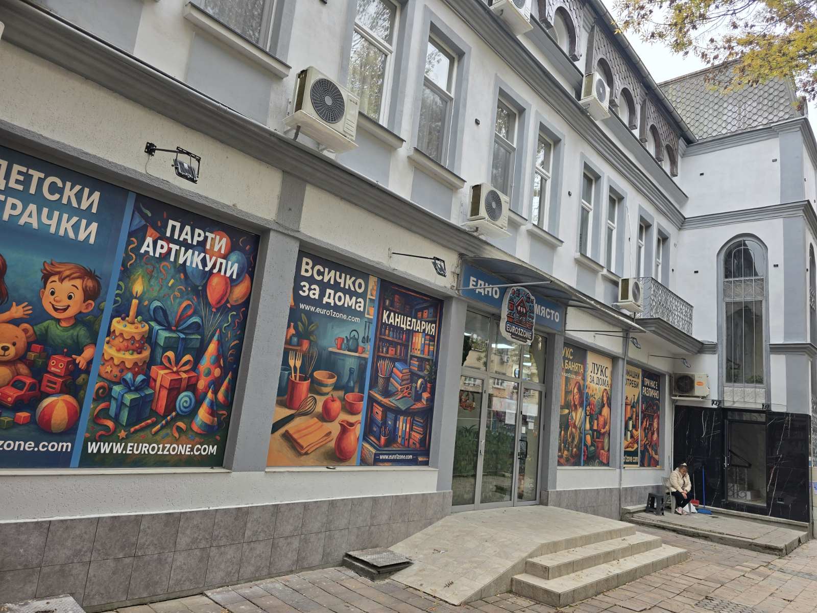 storefront with large colorful posters in Cyrillic advertising gifts and toys on a gray building facade with windows and air conditioners across the upper story