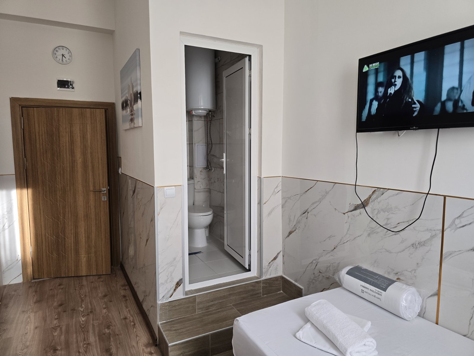 Bed with white linens and rolled towels in foreground, TV on wall to the right, and a small bathroom with glass door opposite, wooden door left, marble wainscoting and wood-look flooring visible.