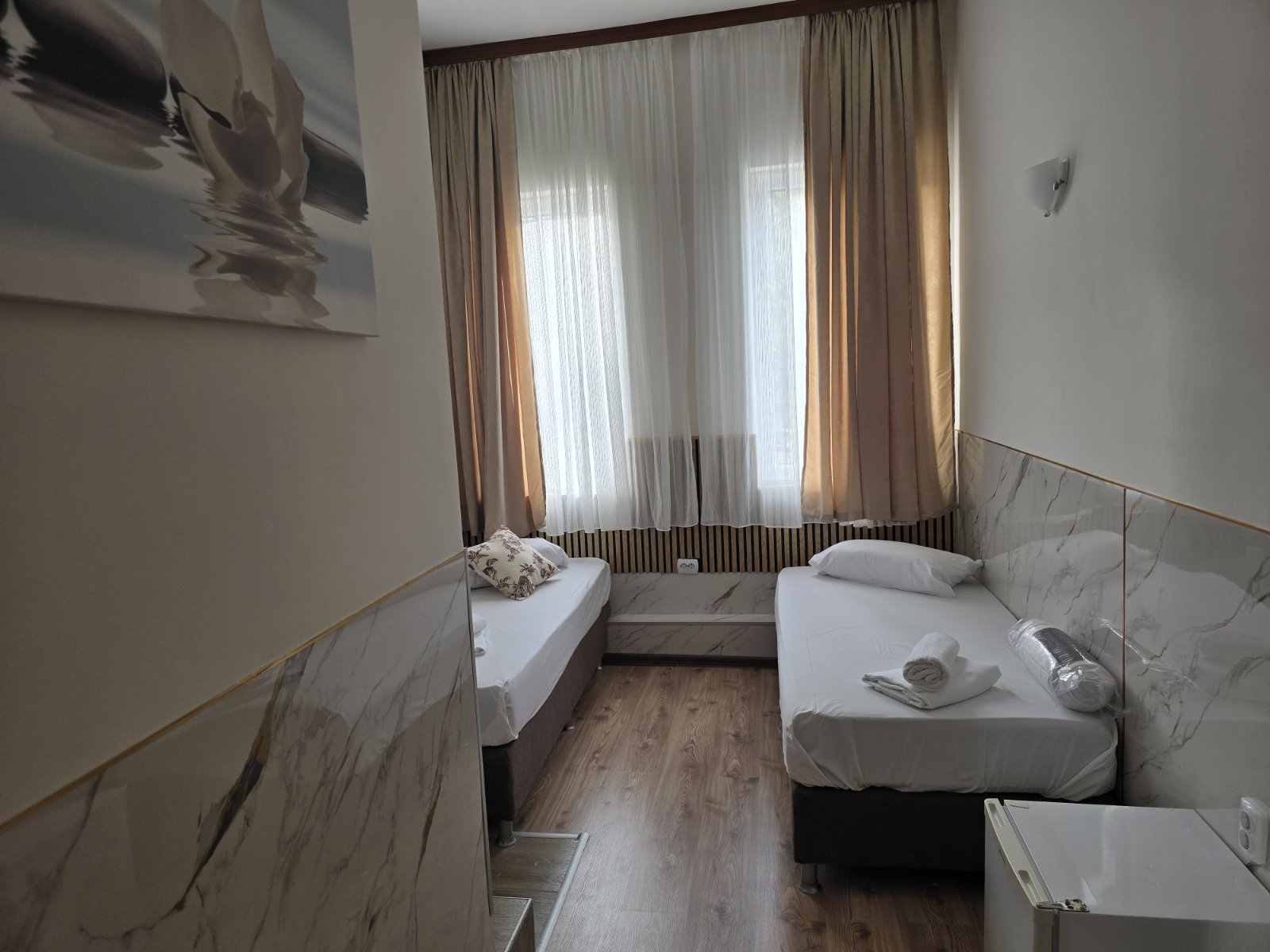 Twin bedroom with two single beds, a large window with sheer white and beige curtains, and marble-style wall panels.