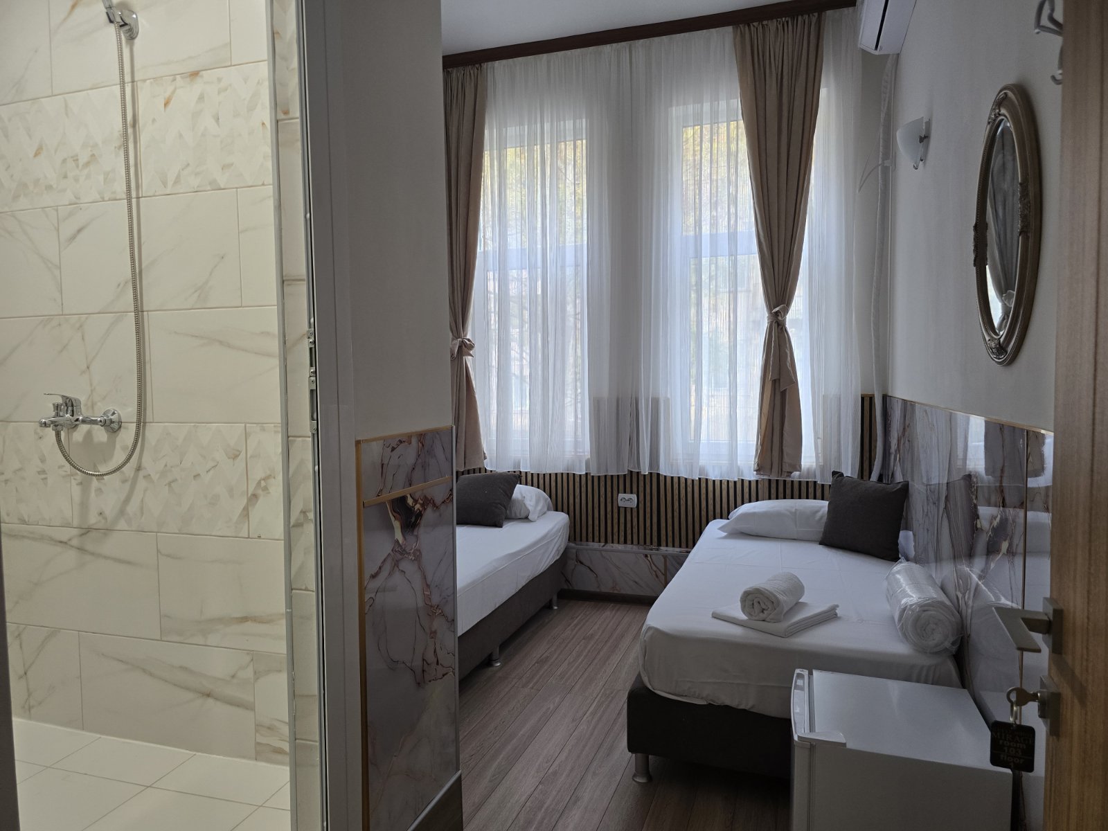 Hotel room with two single beds, sheer curtains, and a window view; bathroom/shower area visible on the left.