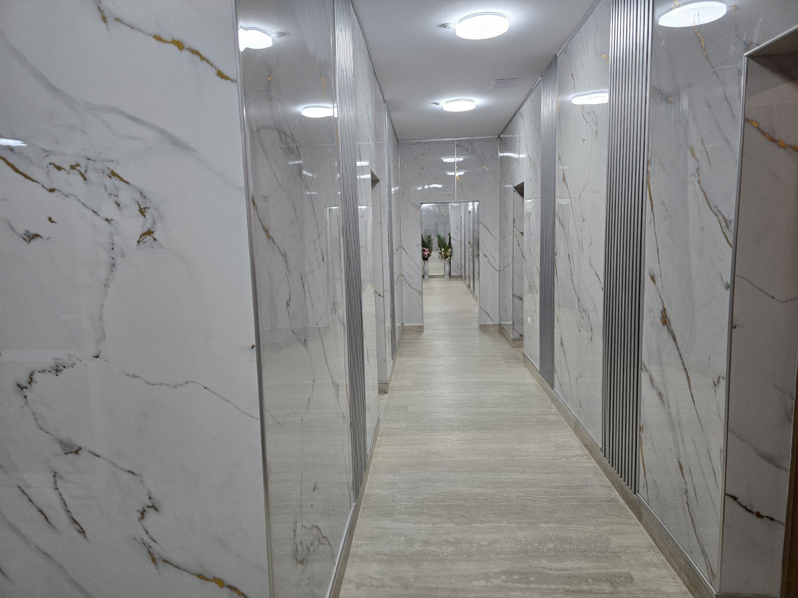 Long marble-walled corridor with mirrored panels, fluorescent ceiling lights, leading to a doorway with potted plants at the end