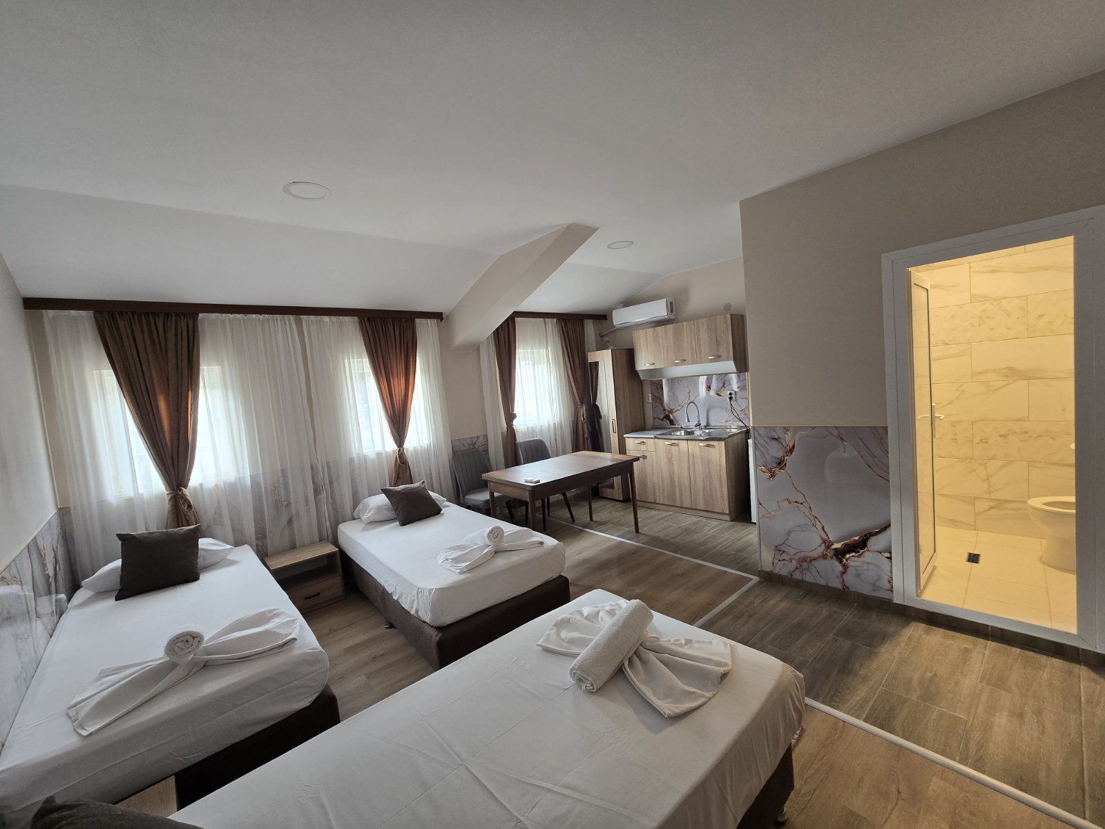Hotel room with three single beds, brown drapes, sheer white curtains, a small table, and kitchenette area; bathroom door on the right.