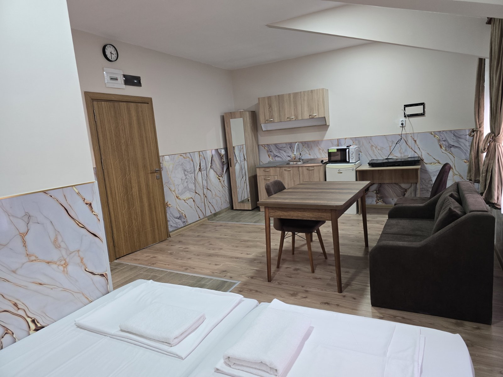Hotel room with a wooden dining table, two chairs, and a brown sofa, kitchenette along the marble-walled wall in the background.