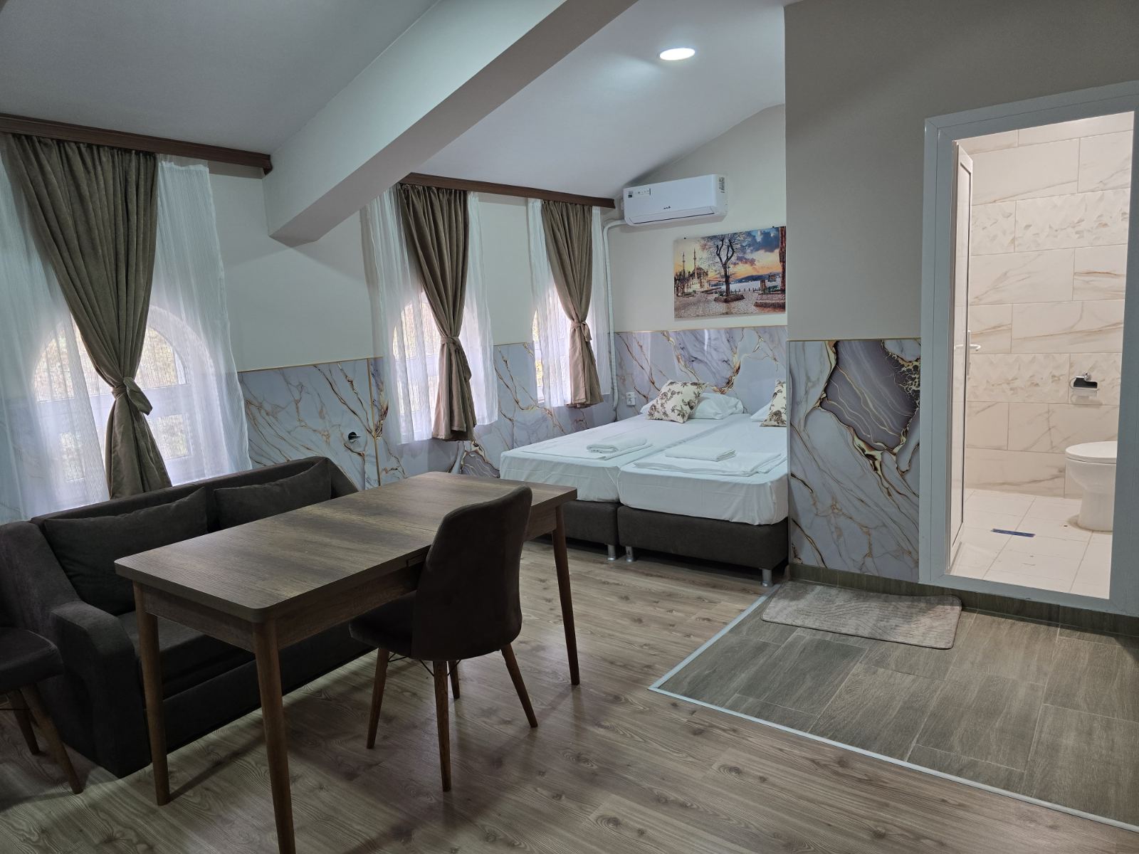 Hotel suite with a seating area, wooden dining table and sofa, marble wall panels, and a bed by windows with beige curtains.